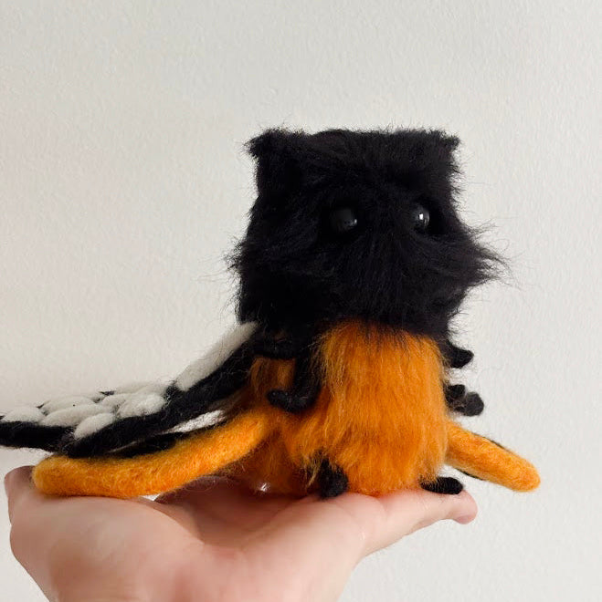 A small handmade felt creature with a black furry head and body, an orange belly and legs, and white wing-like flaps, being held in a person's hand against a plain light wall.