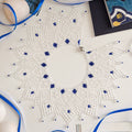 A white crocheted or lace starburst wall hanging with blue bead accents, displayed on a white surface with blue ribbons and decorative props around it.
