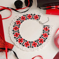 A handmade crocheted collar necklace with a white lace base and red and black floral patterns, displayed on a white surface with craft supplies like ribbons, pencils, and spools.