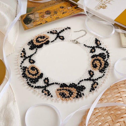 A handmade beaded collar necklace with black and beige rose motifs on a white mesh base, styled on a light surface with white ribbons, a book, and a woven straw hat.