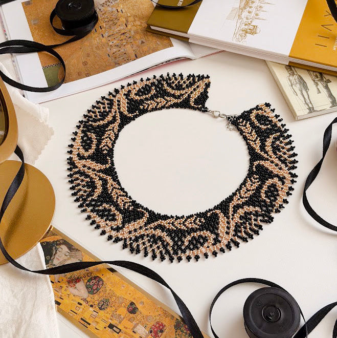 A beaded collar necklace with a black and beige damask pattern, styled on a white surface with books, ribbons, and a camera.