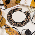 A beaded collar necklace with a black and beige damask pattern, styled on a white surface with books, ribbons, and a camera.