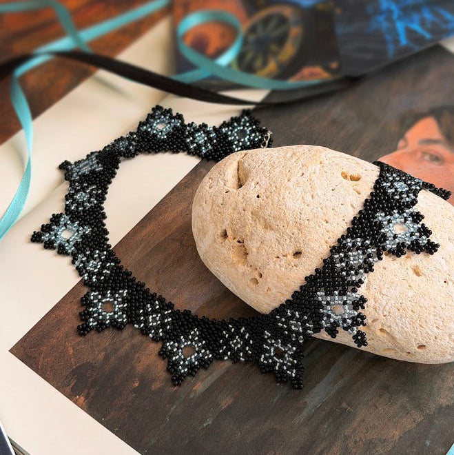 A black and white beaded necklace with a gothic lace-like border, displayed on a rustic wooden surface next to a textured stone prop.